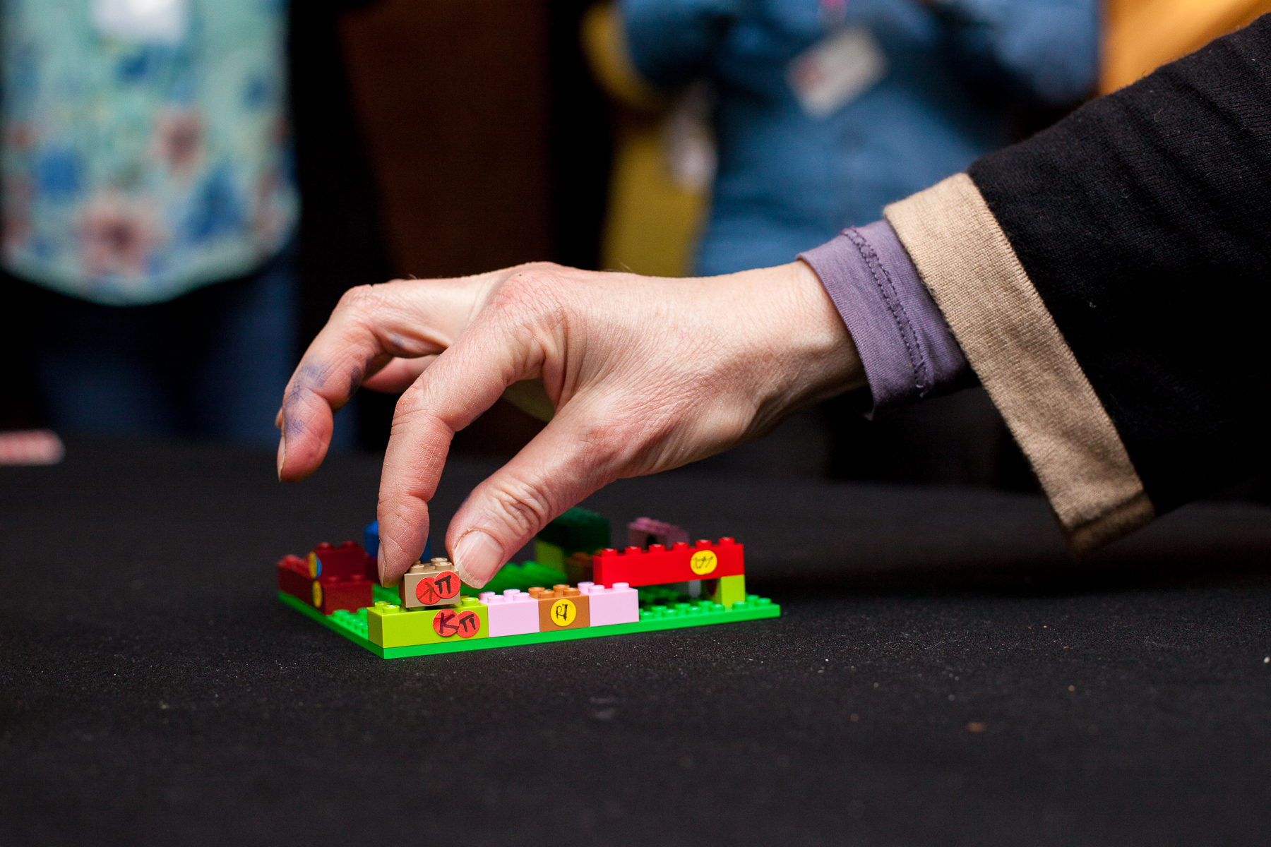 Close-up of a hand placing a small plastic building block onto a green baseplate on a dark table, indicating a collaborative hands-on workshop activity. The layout includes multiple colorful blocks and several small round stickers with handwritten markings that are not legible. The background is out of focus with people and room context, giving a focused and playful tone.