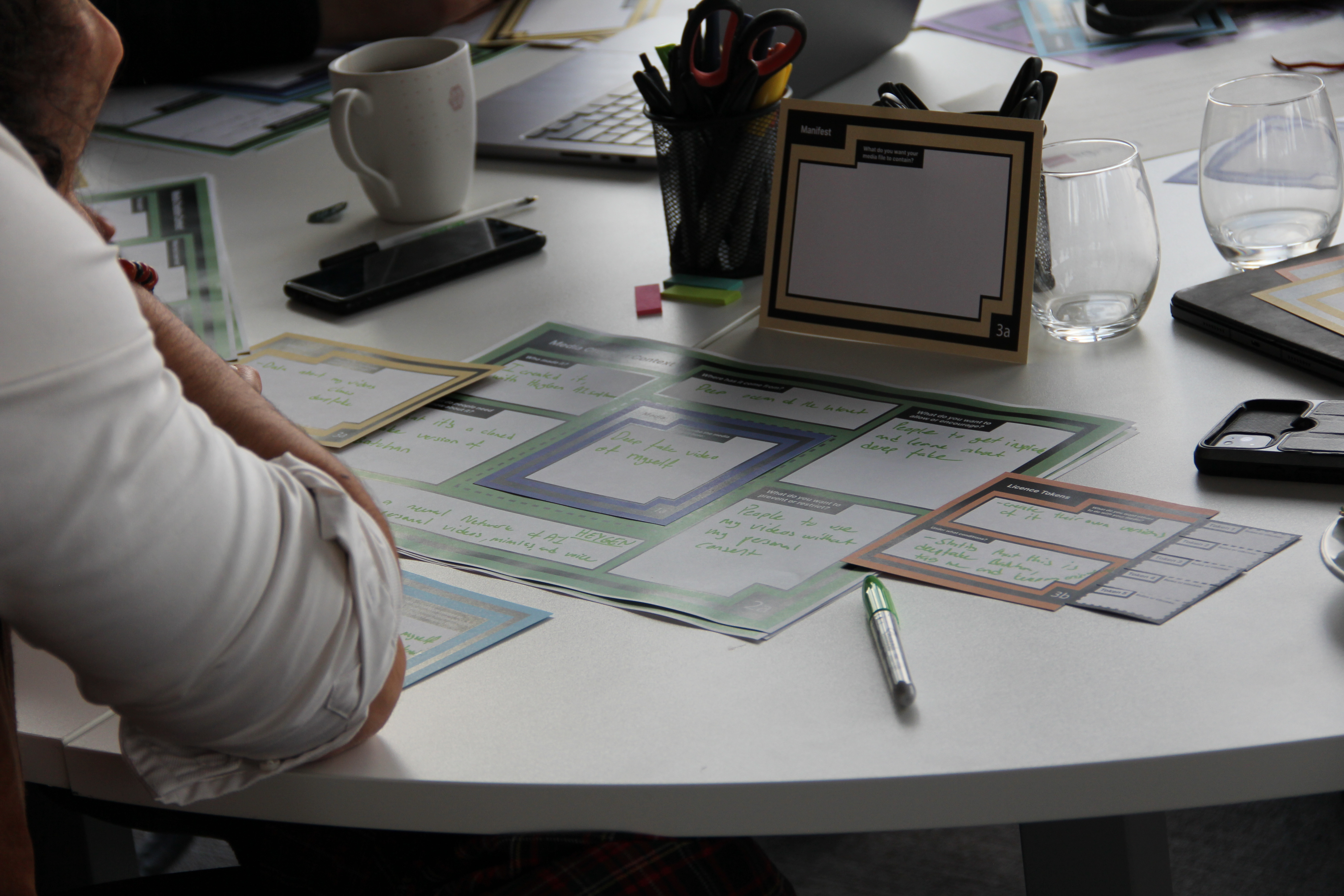 Printed workshop materials and activity worksheets spread across a round white table with a participant looking at the papers; a small folded activity card reads Manifest 3a; handwritten notes on the worksheets are present but not fully legible; the scene shows a focused, collaborative workshop environment.