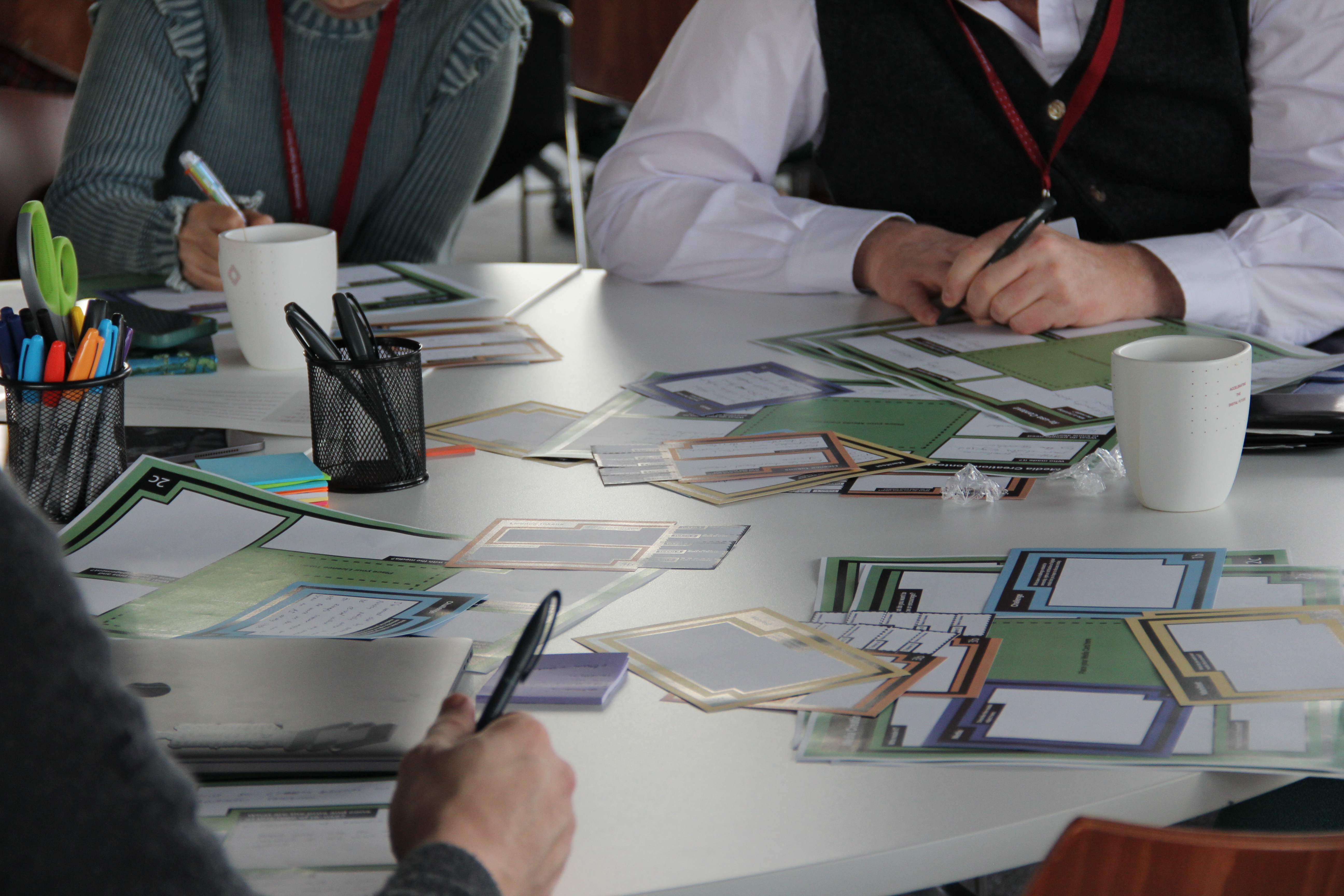Workshop participants seated around a table writing on colourful worksheets and sticky notes with pens; small text on the worksheets is not legible. Indoor workshop setting with a focused, collaborative, playful atmosphere.